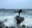 Ein Karateka tritt zur Seite und steht dabei auf einem Stein am Strand, um den herum Wellen aufspritzen, dahinter das Meer