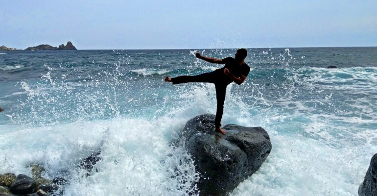 Ein Karateka tritt zur Seite und steht dabei auf einem Stein am Strand, um den herum Wellen aufspritzen, dahinter das Meer