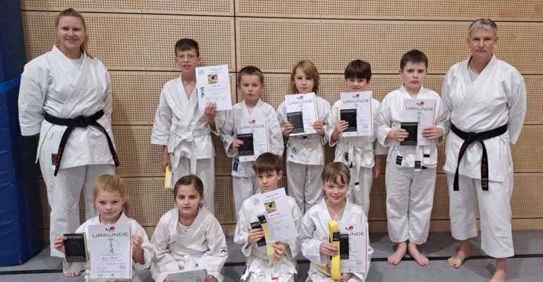 Karate-Kinder mit den Prüfungsurkunden in der Halle, gemeinsam mit den Trainerinnen Sabrina (links) und Susanne (rechts).